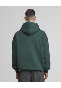 Hettegenser i mørkegrønn, med avslappet passform, myk tekstur og ribbestrikkede mansjetter. Bakfra viser den et minimalistisk design uten synlige logoer.