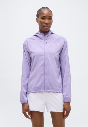 Woman wearing a light purple hooded zip-up jacket and white shorts, standing with a neutral expression against a plain background.