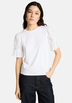 Femme portant un chemisier blanc à manches courtes et bouffantes avec œillets, et un jean bleu foncé, debout avec une main dans la poche sur un fond uni.