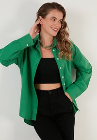 Green button-up shirt with long sleeves, cotton material, side slits, worn over a black crop top, paired with black pants.