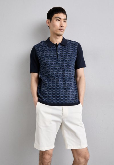 Polo majica marinastolbe barve z teksturiranim geometrijskim vzorcem, kratkimi rokavi in temnim ovratnikom. V kombinaciji s svetlo bež kratkimi hlačami.