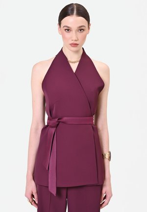 Femme aux cheveux foncés portant un haut prune sans manches à col halter avec une ceinture nouée à la taille et un pantalon assorti sur un fond uni.