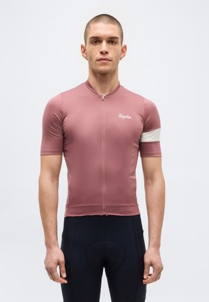 Moški, ki nosi oprijeto prašno roza kolesarsko majico z belo trakico na roki in črne oblazinjene kolesarske hlače, stoji ob enobarvnem ozadju.