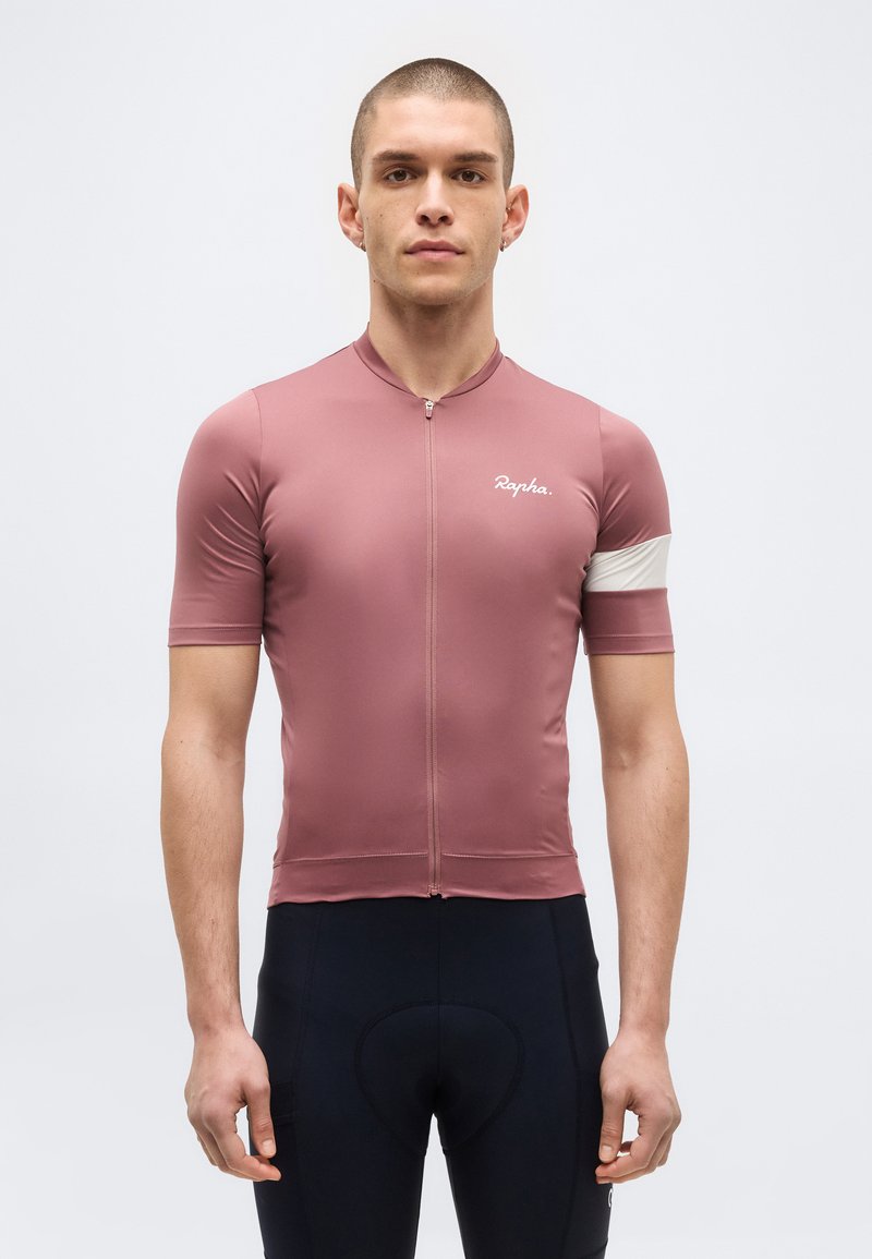 Άνδρας που φοράει εφαρμοστή σκουρόροζ ποδηλατική φανέλα με λευκή ταινία στο μπράτσο και μαύρο παντελόνι ποδηλασίας με επενδύσεις, στέκεται μπροστά σε απλό φόντο.