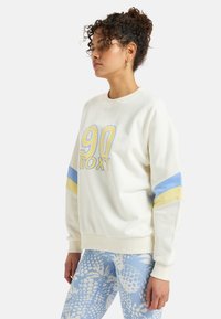 Cream-colored sweatshirt with blue and yellow stripes, featuring a large "90 Roxy" logo. Soft fabric with a relaxed fit and ribbed cuffs.