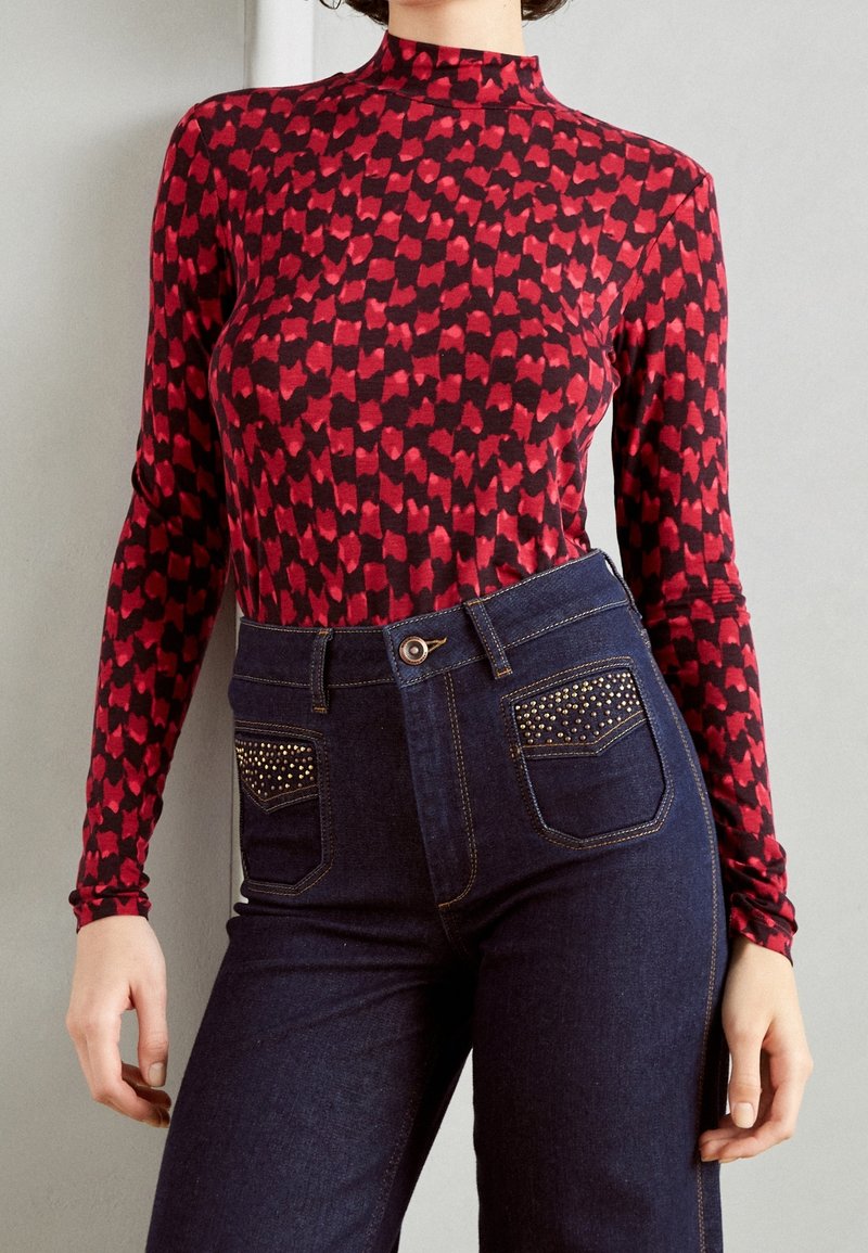 Top a maniche lunghe rosso e nero con un collo alto, abbinato a jeans in denim scuro a vita alta con dettagli di borchie sulle tasche.