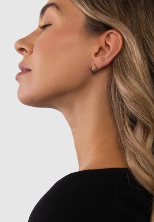 Frau mit geschlossenen Augen, nach links blickend, trägt sonnenförmige Goldohrringe mit einem kleinen Stein in der Mitte, vor einem einfachen Hintergrund.