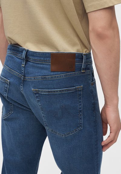 Vue de dos d'une personne portant un jean bleu avec un patch en cuir marron de la marque et une chemise beige rentrée.