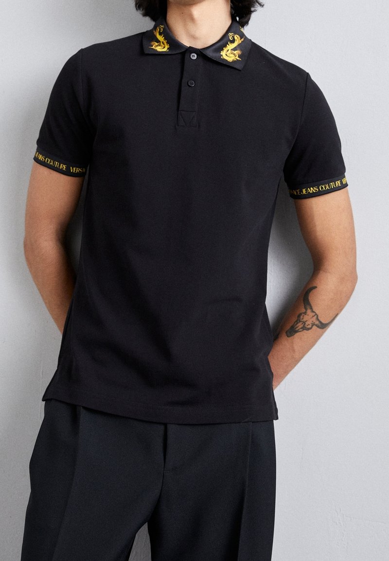 Homme portant un polo noir avec une broderie de dragon en or sur le col et un texte doré "Versace Jeans Couture" sur les poignets des manches.