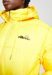 Gros plan sur une personne portant une doudoune jaune vif avec une capuche et un logo "ellesse" noir sur la poitrine.