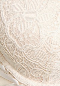 Spitzen-BH in sanftem Beige mit floralen Mustern, glatter Textur und Satinbandakzenten an der Seite, die das Design hervorheben.