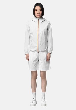 Kobieta stoi w białej kurtce z kapturem na zamek, białych szortach do kolan, białych skarpetach i białych sznurowanych botkach na białym tle.