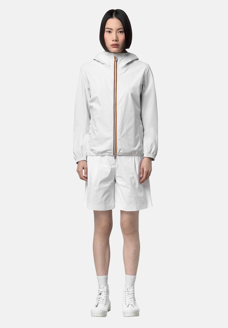 Žena stojaca oblečená v bielej mikine na zips s kapucňou, bielych šortkách po kolená, bielych ponožkách a bielych šnurovacích topánkach na bielom pozadí.