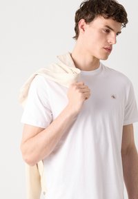 Nuori mies valkoisessa t-paidassa, joka pitää vaaleanruskeaa takkia olallaan ja katsoo alas ja sivulle yksiväristä taustaa vasten.