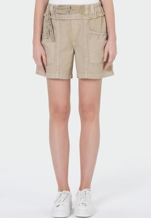 Short beige taille haute avec grandes poches devant, taille ceinturée, porté avec des baskets blanches sur une personne debout.