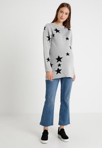 Sweater cinza com padrões de estrelas pretas, mangas longas e um corte descontraído, combinado com jeans azuis de perna larga e sapatos pretos tipo slip-on.