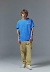 T-shirt en coton bleu à manches courtes, avec un logo sur la poitrine, associé à un pantalon cargo beige et des baskets blanches. Coupe simple et décontractée.