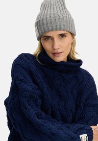 Granatowy sweter z grubym splotem i wysokim kołnierzem oraz prążkowanymi brzegami, zestawiony z jasnoszarym, prążkowanym beanie z metką na mankiecie.
