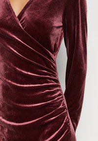 Burgunderröd sammetsklänning med långa ärmar, med en inslagsdesign och veckad textur längs sidan, som ger en figurnära silhuett.