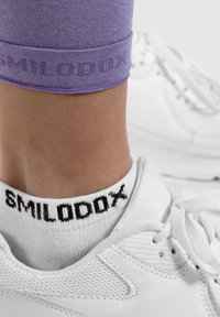 Witte sneaker met een textuur, gecombineerd met enkelhoogte witte sokken met "SMILODOX" in het zwart, en paarse leggings met logo aan de zoom.