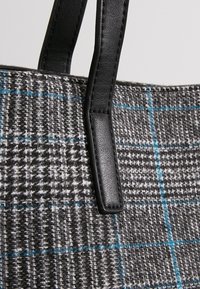 Sac fourre-tout à carreaux gris et noirs avec des accents bleus ; dispose de poignées en cuir noir et d'un tissu tissé texturé.