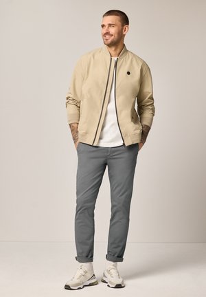 Mann steht mit Händen in den Taschen, trägt eine beige Bomberjacke, weißes T-Shirt, graue Hose und weiße Sneakers vor einfarbigem Hintergrund.