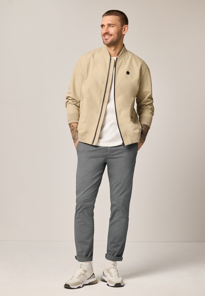 Homme debout les mains dans les poches, portant une veste bomber beige, un t-shirt blanc, un pantalon gris et des baskets blanches sur fond uni.