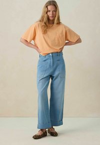 T-shirt en coton orange clair avec une poche poitrine, associé à un jean léger à taille haute et jambes larges bleu clair, avec de grandes poches arrière et des ourlets roulés.