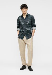 Νέος άνδρας με σκούρο πουκάμισο με σχέδια και κουμπιά, με τα μανίκια γυρισμένα, μπεζ παντελόνι και μαύρα παπούτσια, στέκεται με τα χέρια στις τσέπες.