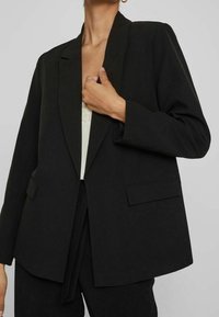 Černý sako s klopami s výstřihem do V, s jedním knoflíkem a dvěma předními kapsami. Hladká tkanina a volný střih, nošený přes lehký top.