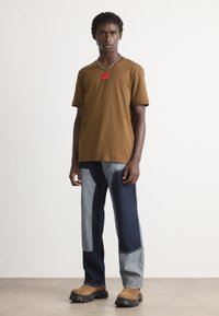 Ung mann iført brun t-skjorte med rød logo, lappede denimjeans, brune støvler og sølvkjede, stående foran en ensfarget bakgrunn.