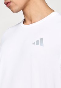 Λευκό αθλητικό μπλουζάκι με ένα διακριτικό ασημί λογότυπο. Το ύφασμα έχει ομαλή υφή με λεπτές οριζόντιες ραβδώσεις.