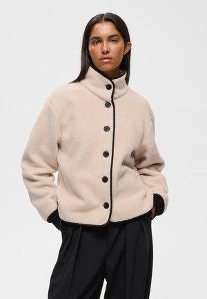 Femme aux longs cheveux foncés portant une veste polaire beige avec des boutons noirs et un pantalon noir, debout les mains dans les poches devant un fond uni.