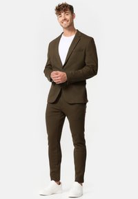 Completo verde oliva con giacca su misura e pantaloni slim-fit, abbinato a una t-shirt bianca e sneaker bianche. Tessuto liscio, linee pulite.