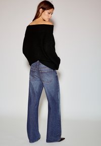 Zwarte trui met blote schouders, gecombineerd met blauw denim jeans met wijde pijpen. De jeans heeft vijf zakken en subtiele stiksel details.