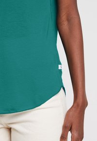 T-shirt turchese con orlo rotondo e dettagli con spacco laterale, realizzato in tessuto morbido. Presenta un piccolo tag sul lato sinistro vicino all'orlo.