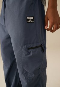 Pantaloni cargo blu navy in tessuto leggero con una tasca con cerniera, caratterizzati da una toppa con la scritta "SUBSCRIBE LDN" sulla coscia superiore.