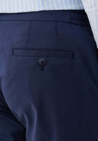 Tasca posteriore dei pantaloni blu navy con chiusura a bottone, indossati con una camicia a righe azzurre e bianche.