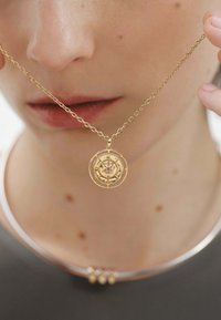 Collar de oro con un colgante circular que presenta diseños intrincados y una piedra central. La cadena tiene segmentos ovalados enlazados con un acabado brillante.