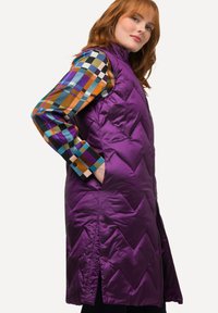 Lila Steppweste mit einem Rautenmuster, ausgestattet mit einem hohen Kragen, seitlichen Taschen und einem Reißverschluss. Sichtbare Ärmelbündchen eines mehrfarbigen Karomusters.