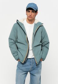 Teal boblejakke med en hvit fleece-fôrkrage, detaljer i gul og rød glidelås, båret over en hvit t-skjorte og lyseblå jeans.