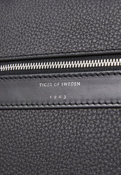 Détail d'un sac en cuir noir texturé mettant en avant une fermeture éclair en argent, un logo embossé "TIGER OF SWEDEN" et l'année "1903".