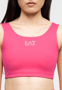Γυναίκα που φοράει ένα φωτεινό ροζ αμάνικο crop top με λευκό λογότυπο EA7 Emporio Armani στο κέντρο του στήθους.