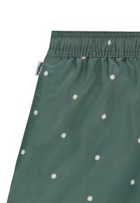 Shorts en tissu vert avec une taille élastique, arborant un motif de marguerites éparpillées en blanc et jaune. Étiquette visible sur le côté de la taille.