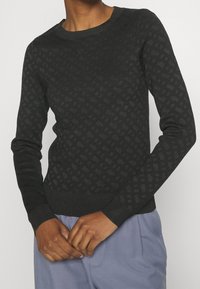 Pull noir avec un motif en losange texturé, col rond côtelé et poignets côtelés. Tissu apparemment doux, silhouette ajustée, associé à un pantalon clair.