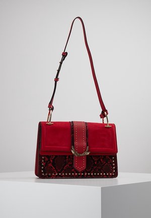 Sac à bandoulière rouge avec motif peau de serpent, clous dorés, boucle décorative dorée et sangle réglable sur une surface d'exposition blanche.