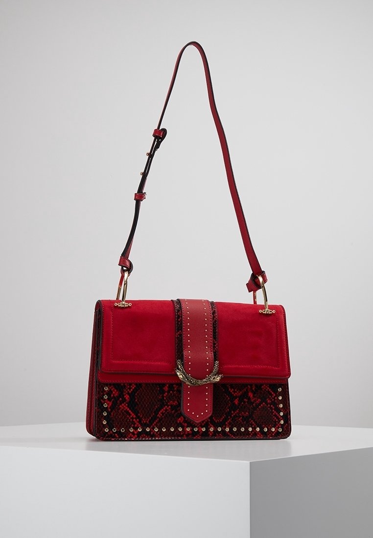 Sac à bandoulière rouge avec motif peau de serpent, clous dorés, boucle décorative dorée et sangle réglable sur une surface d'exposition blanche.