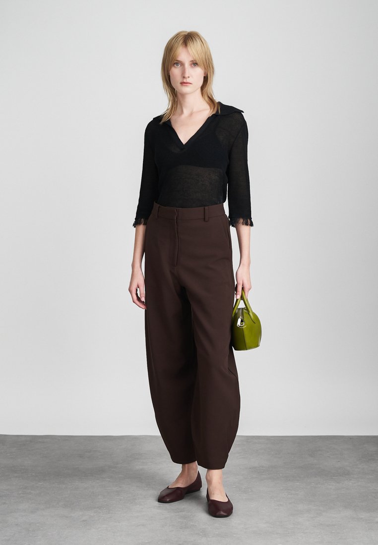 Black sheer top with a collar, brown wide-leg trousers, and burgundy flats. Holding a small, structured green handbag with silver hardware.
