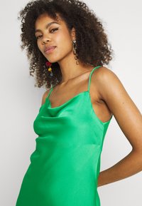 Vestido de seda verde com decote em cowl e alças finas, apresentando uma textura suave e uma silhueta justa, combinado com brincos coloridos.