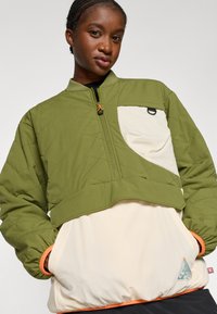 Chaqueta pullover acolchada verde con una sección inferior color crema, que presenta un acento naranja en el dobladillo, un cierre de cremallera y un bolsillo lateral.